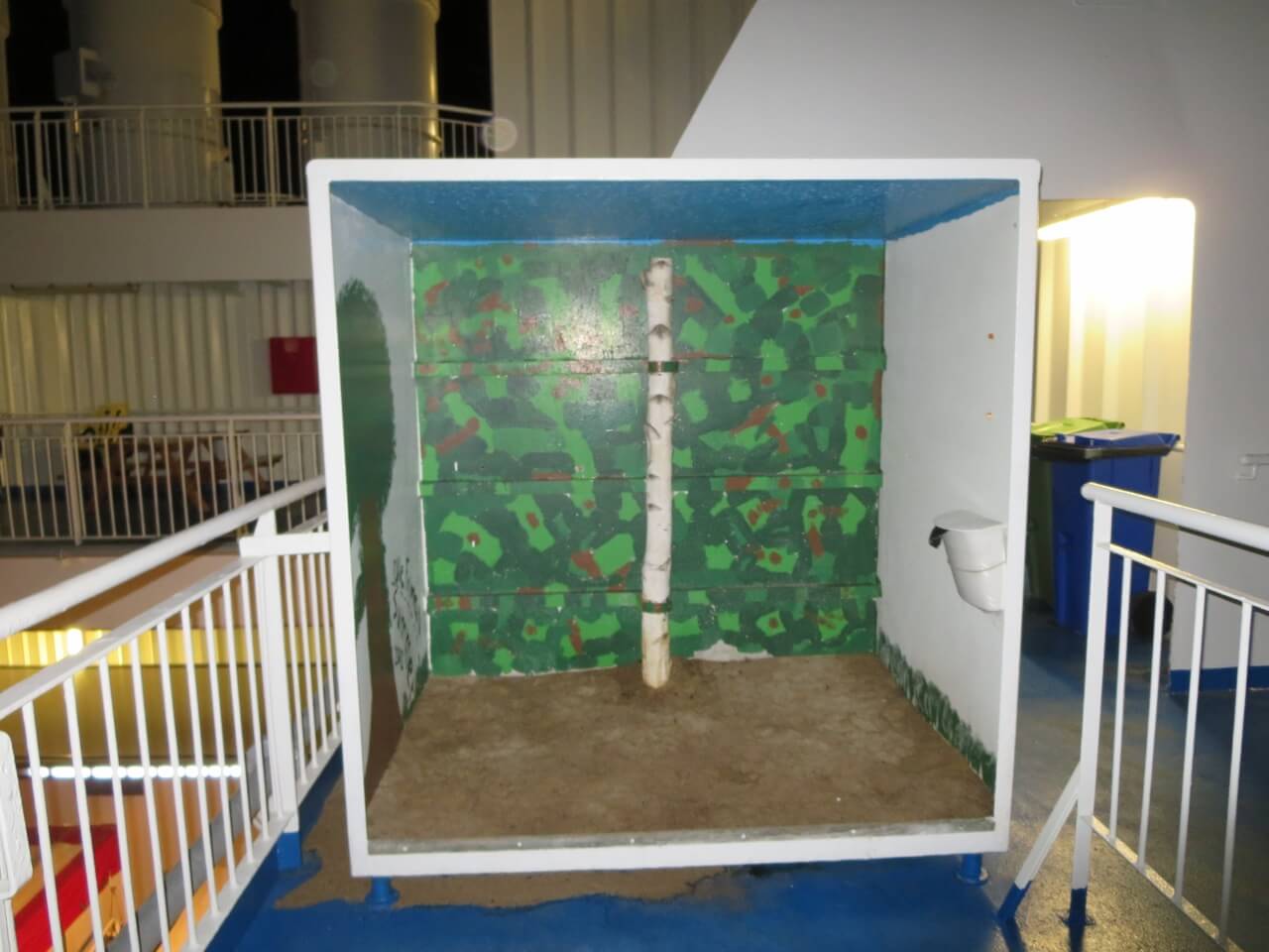 Eine große nach oben geöffnete Kiste mit Sandboden und einem gemalten Wald an den Wänden