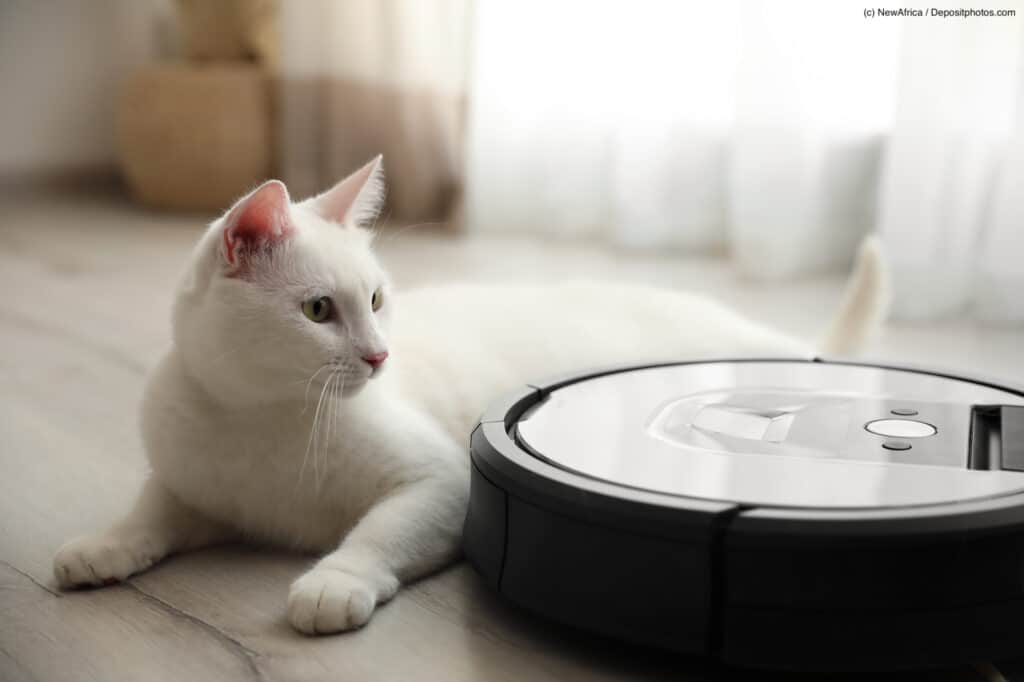 Weiße Katze liegt auf Parket und schaut interessiert zu einem Staubsaugerroboter neben ihr. Bildbeschreibung lautet: Saug- und Wischroboter helfen dir als Allergiker deine Wohnung sauber zu halten und nicht zu viel Staub und Haare aufzuwirbeln.