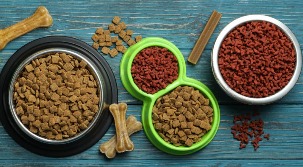 Verschieden große Futternäpfe, gefüllt mit Trockenfutter stehen auf einem Tisch, darum Kauknochen. Beschriftung lautet: In manchen Fällen sind viele kleinere Mahlzeiten besser als eine große.
